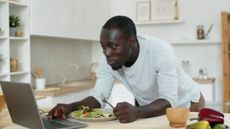 Hombre-Afroamericano-Comiendo-Ensalada-Y-Videollamadas-En-Una-Laptop-En-La-Cocina